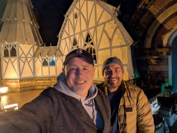 Two men smiling for a selfie in front of illuminated buildings at night.